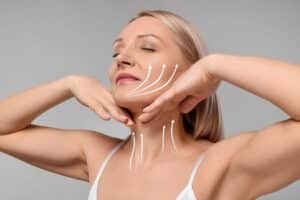 Middle-aged woman gently lifting her chin and jawline with her hands, with illustrative arrows showing lifting effects on the cheeks and neck, representing facelift benefits.